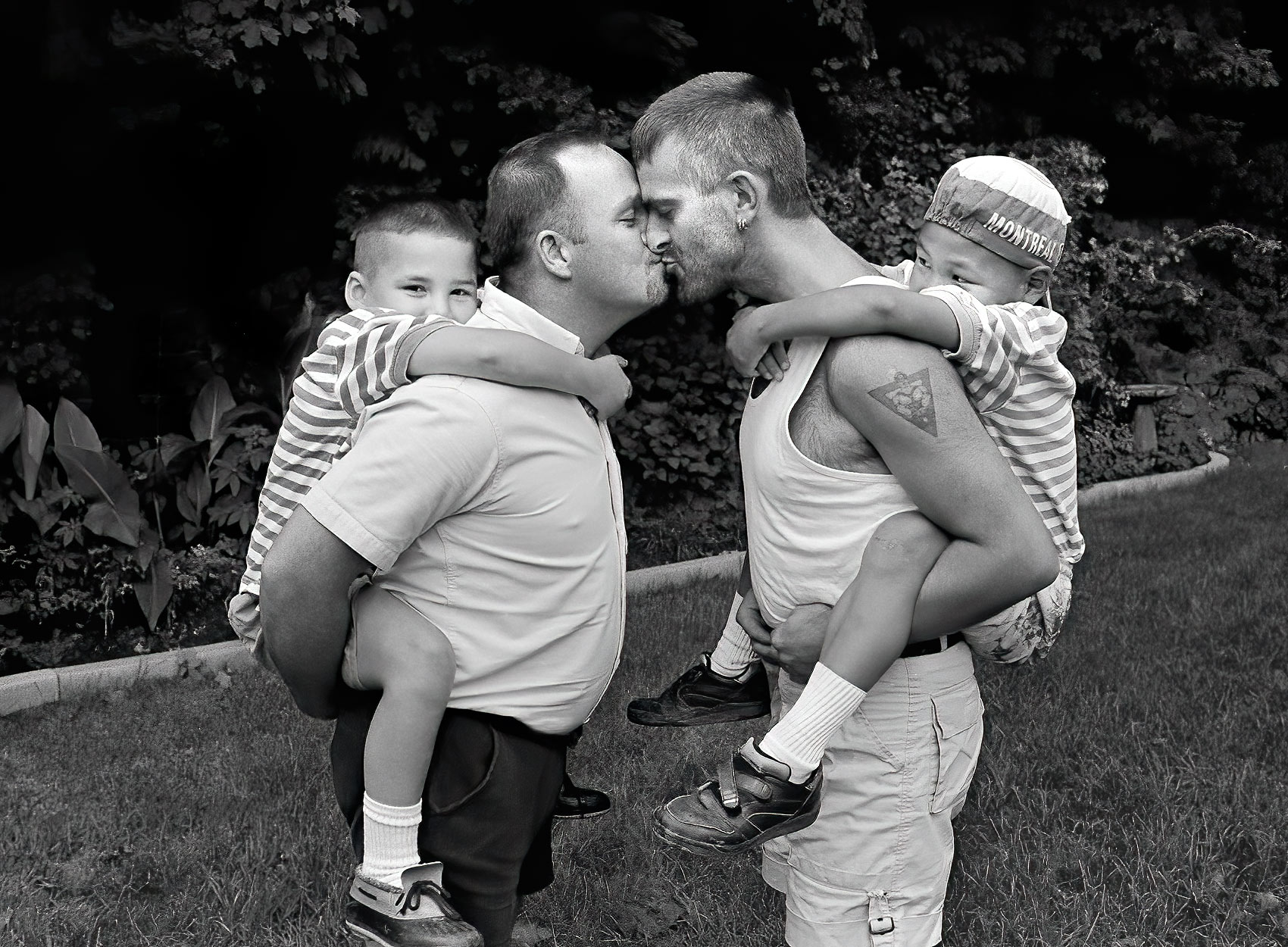 a gay couple piggy back their adopted inuit sons on their backs as they both kiss each other