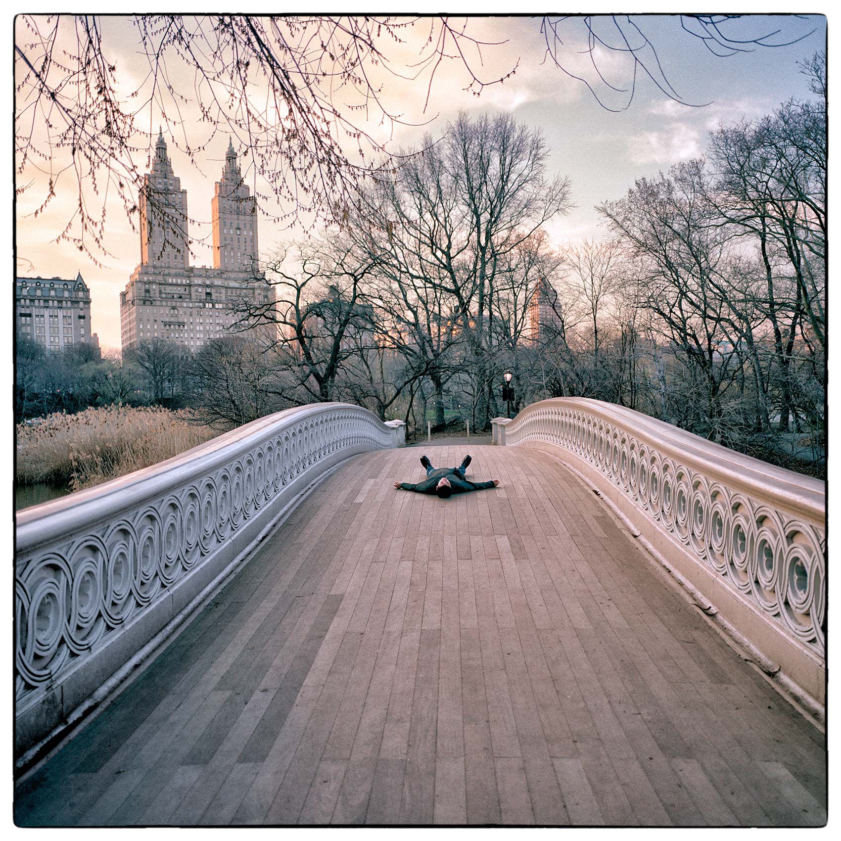 a-man-lays-in-the-middle-of-a-bridge-in-new-yorks-central-park-creating-an-artistic-photo-in-the-late-afternoon