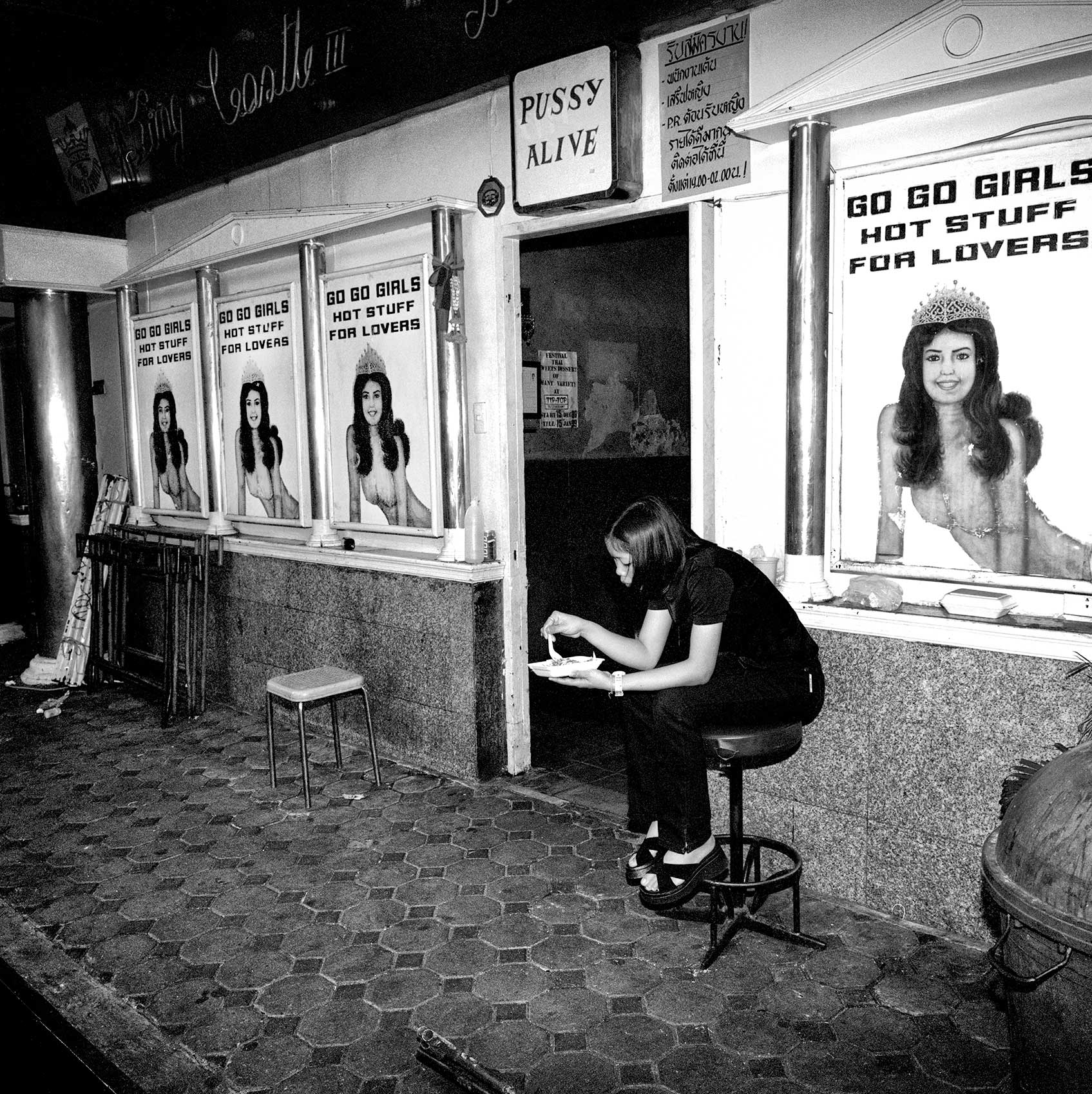 a-woman-eats-food-as-she-works-the-door-of-a-strip-club-in-bangkok-thailand