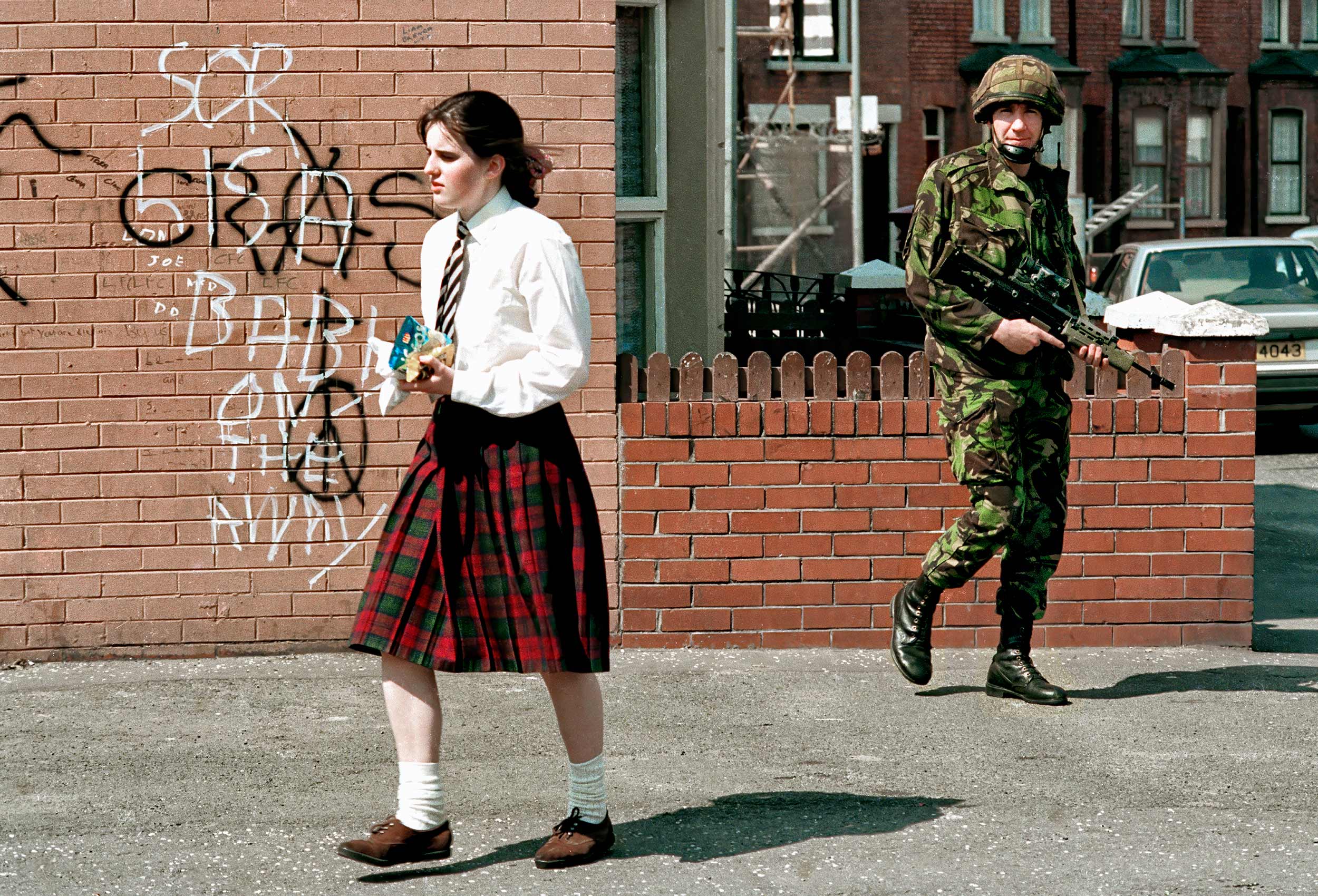 a young school girl walks past a british soldier with a gun in catholic west belfast ireland