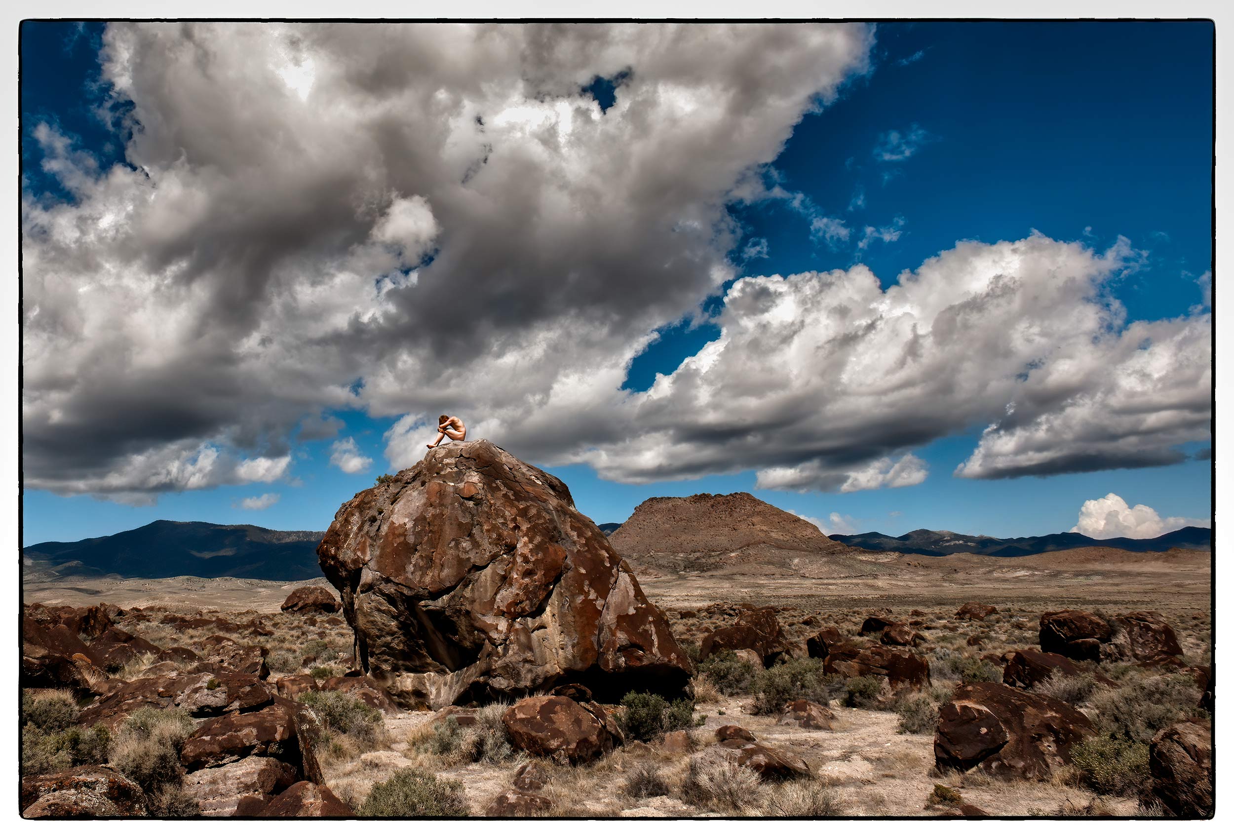 a-young-man-sits-naked-on-top-of-a-giant-boulder-in-the-nevada-desert-surrounded-by-blue-skies-and-clouds