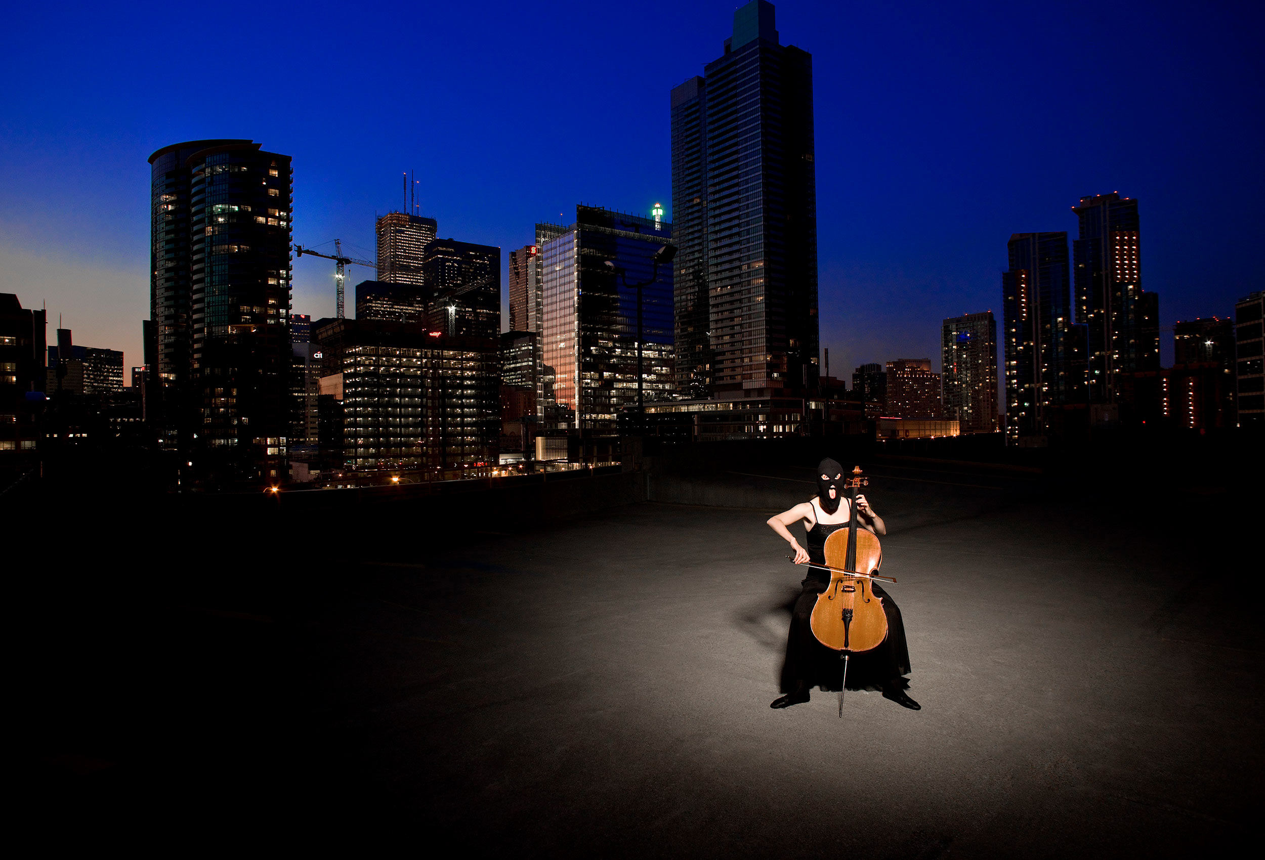 a women wearing a ski mask plays the cello in front of the toronto city skyline